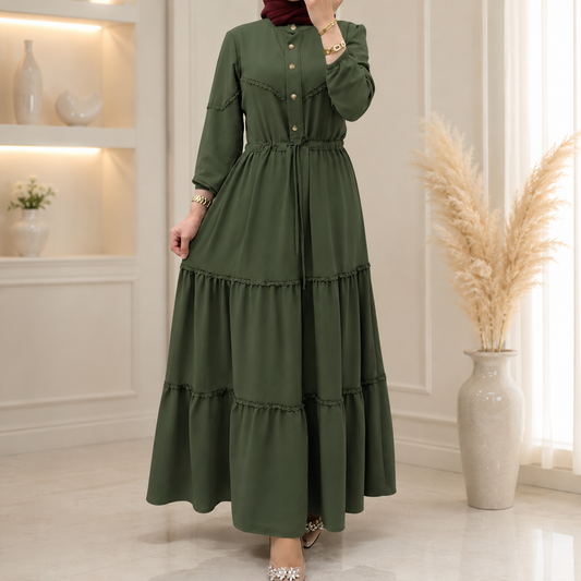 A woman models a long-sleeved olive green maxi dress with tiered ruffles and gold buttons, standing in a bright, modern room with a pampas grass vase.