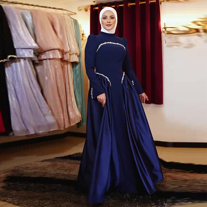 A woman wearing a long-sleeved navy blue maxi gown with silver embellishments and a matching white hijab, standing in a boutique with other dresses hanging in the background.