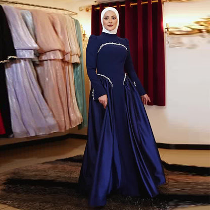 A woman wearing a long-sleeved navy blue maxi gown with silver embellishments and a matching white hijab, standing in a boutique with other dresses hanging in the background.