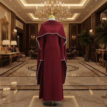A long burgundy abaya dress displayed on a mannequin, featuring wide draped sleeves with white beaded trim and a gold mannequin stand against a white paneled wall.