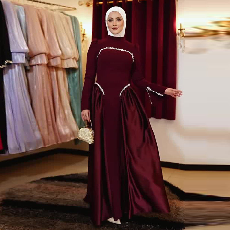A woman wearing a long-sleeved maroon satin modest dress with white bead trim and a matching headscarf, holding a small gold handbag in an indoor showroom with other dresses in the background.