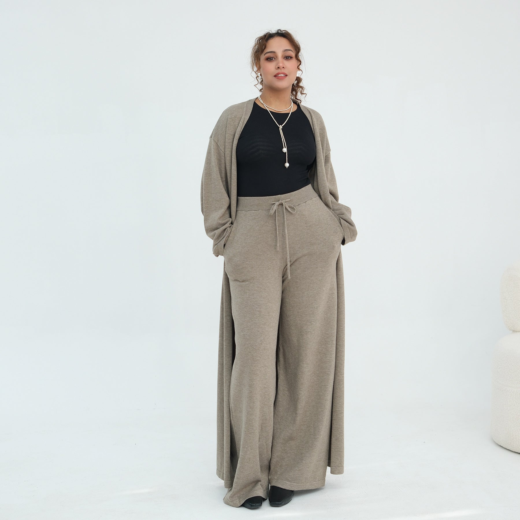 A woman modeling a taupe knit cardigan and matching wide-leg drawstring pants over a black ribbed top, photographed against a clean white studio background.