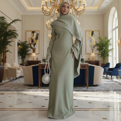 Woman in a long green dress with a hood standing in a room with a lamp and table.