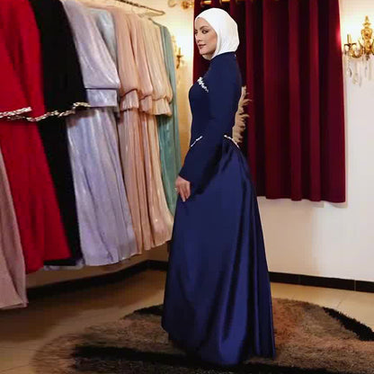 A woman wearing a navy blue long-sleeved maxi dress with a white hijab stands in a boutique showroom. The background features racks of various colorful formal dresses and deep red curtains.