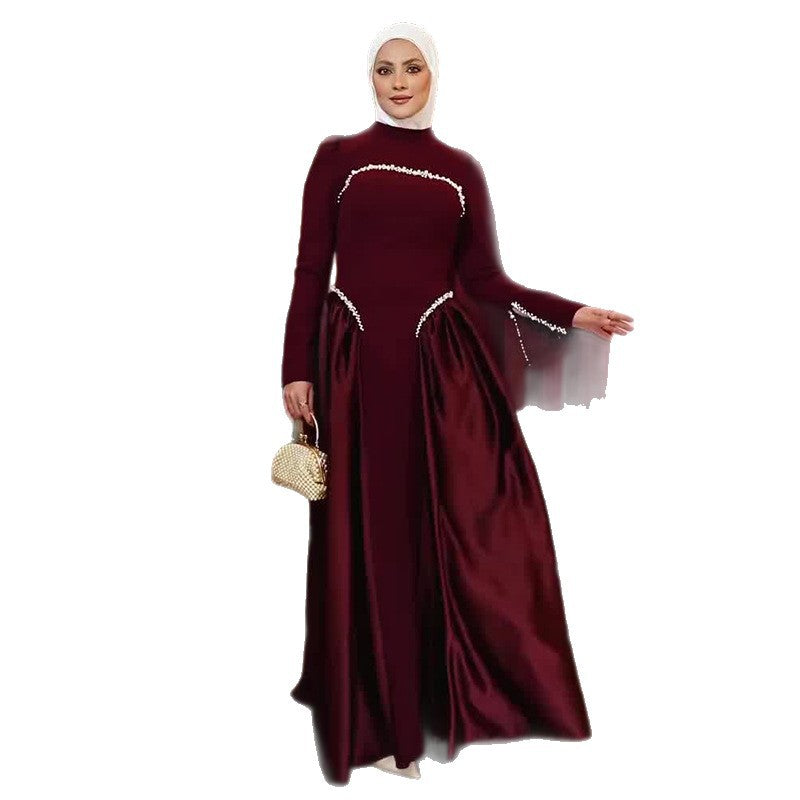 A woman wearing a long-sleeved, floor-length maroon abaya dress with white beaded trim details, a white hijab, and holding a small gold clutch purse, set against a plain white background.