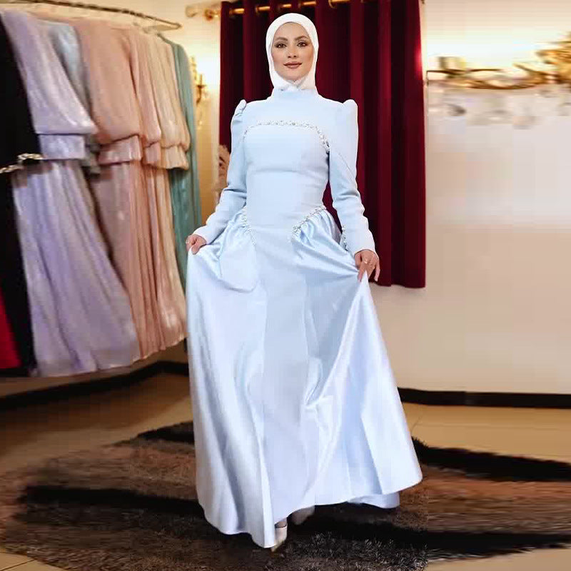 A woman wearing a light blue satin maxi dress with a white hijab stands in a boutique, holding out the skirt to show its drape. The background features other colorful dresses hanging on racks and a dark red curtain.