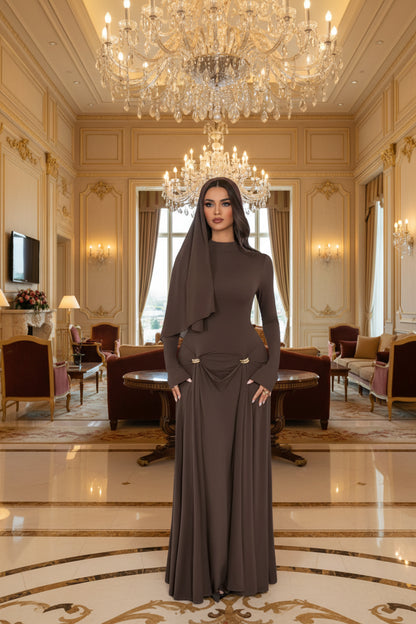 A woman models a long-sleeve, high-neck, floor-length brown dress with a draped waist detail and small gold-tone decorative accents. The setting is a bright, modern room with white paneled doors, a white armchair, and scattered red rose petals on the floor.
