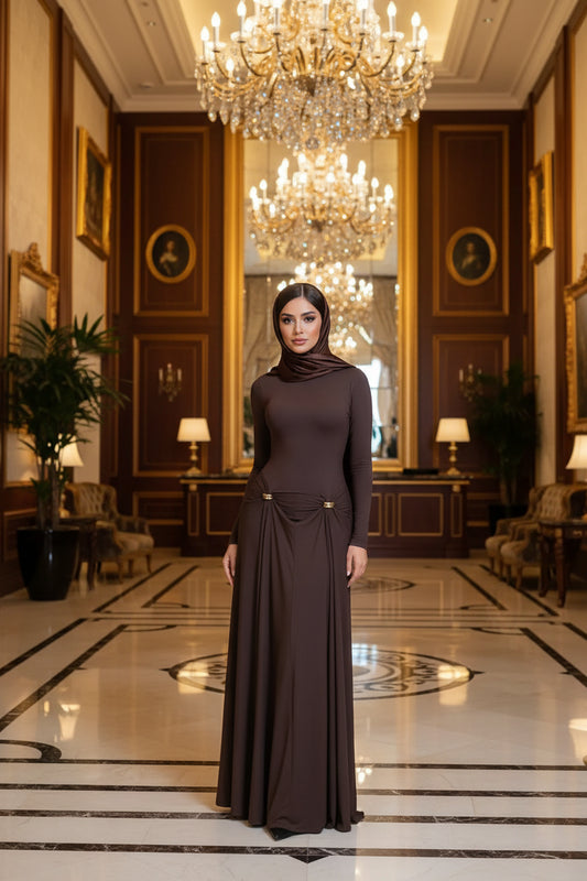 A woman models a long-sleeve, floor-length brown maxi dress with a gathered waist detail accented by two small gold rings, standing in a brightly lit room with a white chair and door.