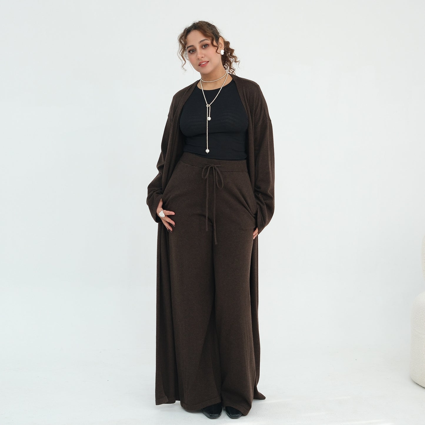 A woman modeling a dark brown two-piece outfit consisting of a long-sleeve cardigan and matching wide-leg drawstring pants, worn over a black top, against a plain white studio background.