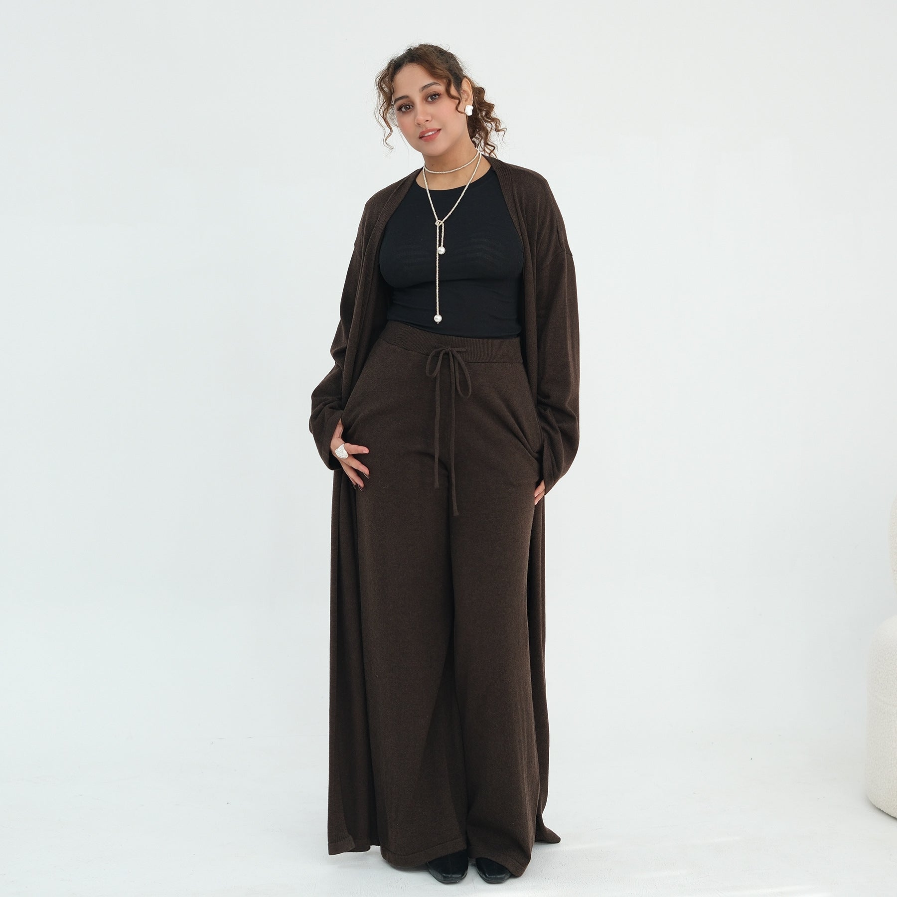 A woman modeling a dark brown two-piece outfit consisting of a long-sleeve cardigan and matching wide-leg drawstring pants, worn over a black top, against a plain white studio background.