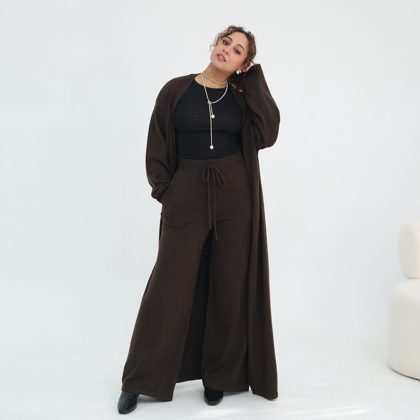 A woman models a dark brown three-piece outfit consisting of a long-sleeve cardigan, a black ribbed top, and matching wide-leg drawstring pants, set against a clean white studio background.