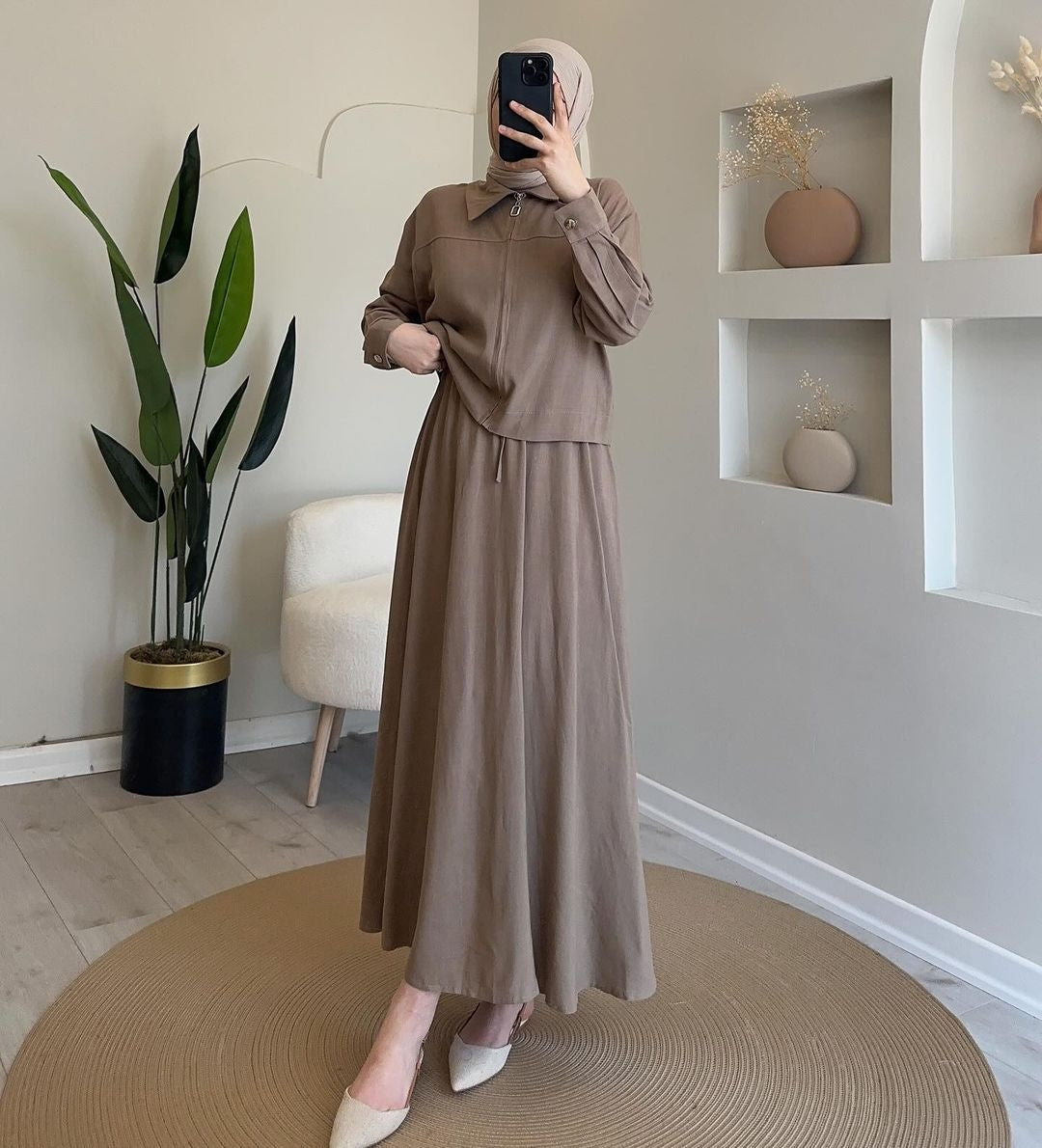 A woman wearing a matching taupe brown two-piece outfit consisting of a long-sleeve collared top and a long maxi skirt, paired with a beige hijab and white pointed-toe flats, posing in a modern neutral-toned room.